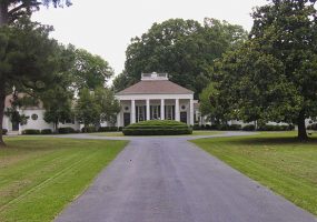 Mansion house with covered porch and single-story wings on estate with paved driveway and trees