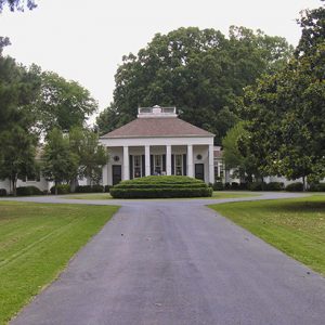 Mansion house with covered porch and single-story wings on estate with paved driveway and trees