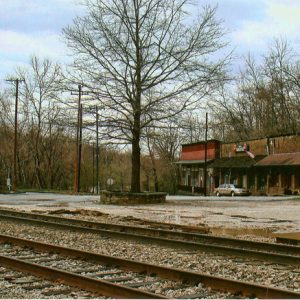 Three brick storefronts with covered porches and covered bridge on street with pair of railroad tracks in the foreground