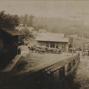 Horse drawn wagons parked outside single-story storefront inside fence across from a multistory building with balcony