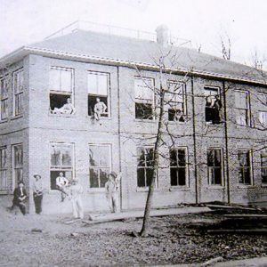 Two-story brick building with people standing outside and sitting in and looking from top floor windows