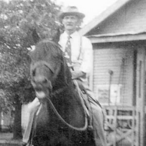 Old white man on horseback with building with covered porch behind him