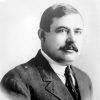 White man with dark hair and mustache in suit jacket and tie