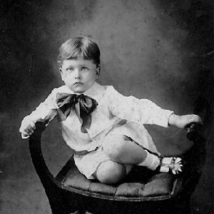 serious white boy in formal dress with large bow tie sitting on a backless chair