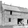 Two-story log cabin with brick chimney with white family out front