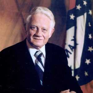 Old white man smiling in suit and tie with Arkansas flag behind him