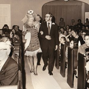 White man in dress and veil with man in suit in crowded church sanctuary