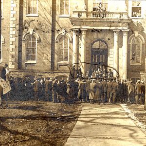 Crowd of white men women and white man on horseback outside multistory building with balcony and arched windows