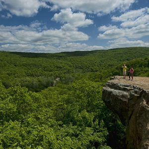 Two men with cameras standing on cliff edge surrounded by tree covered mountains and blue skies