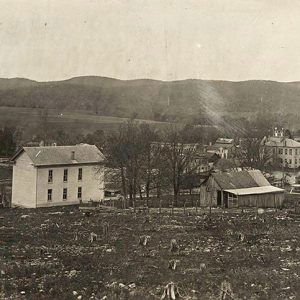 Multistory house and barn in field with multistory town buildings and hills in the background