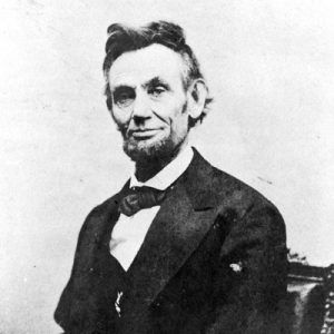 Portrait bearded white man seated at desk smiling disheveled hair suit bow tie