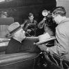 White man in a car talking to a tall white man in a long coat next to a Pullman train car with people standing around them