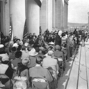 White man in suit speaking at a lectern with crowd of people in chairs behind him