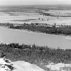 Aerial view of river and fields in the background, white man and woman perched on rock outcropping in foreground