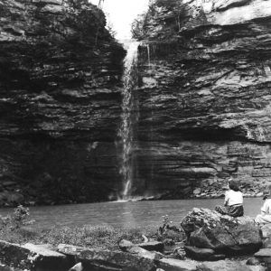 Natural waterfall and pool being observed by two woman sitting on rocks at the edge of the water