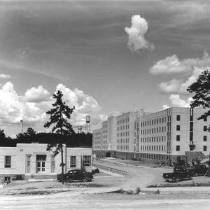 Tall and long building with rows of windows and outbuilding with trees and water towers in the background