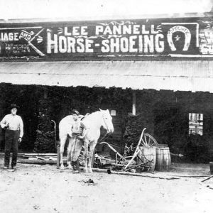 White man and women with horse in front of "Lee Pannell Horse Shoeing" shop