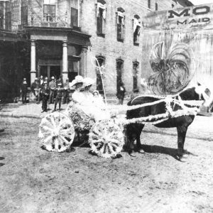 Two white women with little girl in white on a horse drawn buggy with brick building and people in the background