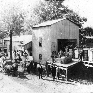 Men posing outside barn on loading dock with bundles near horses, wagons