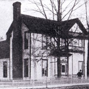 Two story wood frame house with chimney and man in yard behind iron fence