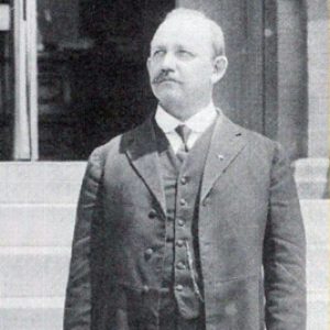 White man on steps looking upward in three piece suit with long jacket holding hat