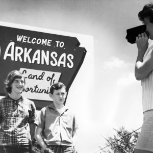 White girl and boy photographed by woman in front of sign "Welcome to Arkansas Land of Opportunity"