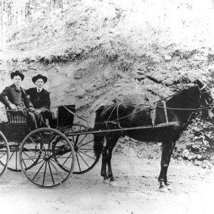 Two men pose in suits, hats, on horse-drawn carriage by dirt back slope