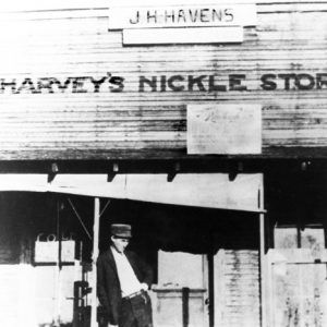 White man in hat hand in pocket leaning on "Harvey's Nickle Store" porch post