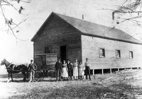White men women pose with horse and carriage by wooden A-frame building, lumber piles