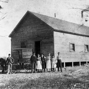 White men women pose with horse and carriage by wooden A-frame building, lumber piles