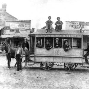 White men pose on horse-drawn trolley labeled "Walnut Ridge & Hoxie, 2" with store, restaurant behind