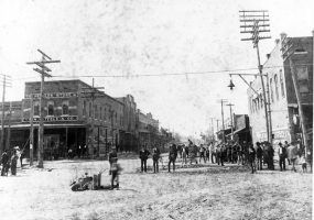 Boy at downtown intersection with equipment, crowd of pedestrians, brick building sign "The Peoples Store"