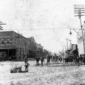 Boy at downtown intersection with equipment, crowd of pedestrians, brick building sign "The Peoples Store"