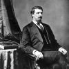 Portrait of white man in suit, tie, seated by curtain, table, and book