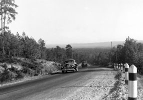 Mountain highway with two cars driving uphill past striped posts on gravel shoulder