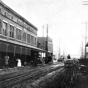 Dirt city street lined with brick architecture, women in dresses below awning, horse drawn carriages, pedestrians