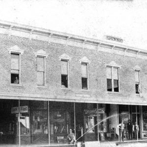 Man sprays water hose outside brick building with sign "Opera Block"  and shops with onlookers in second floor windows