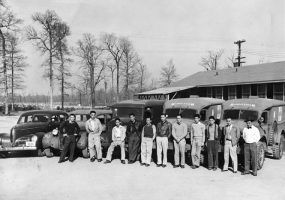 Japanese American men in casual attire pose before cars, ambulances in lot outside wood frame building