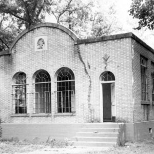 Brick building in woods with large windows arched facade including centered Native American relief portrait