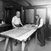 Office two white men examine large sheets paper on sawhorse desk with bricks opposite table