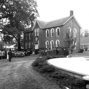 Audience and choir outside two-story brick building with "AC" logo banner reading "1872-1972 100 years"