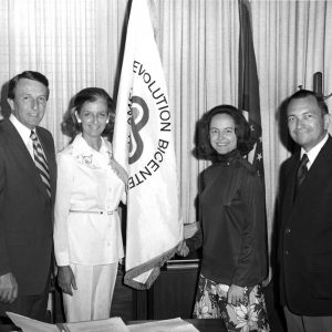 Interior two white men and women pose by flag reading partly "Revolution Bicentennial"