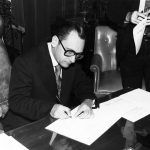 Interior, white man in suit seated signing paper, with white man standing holding form