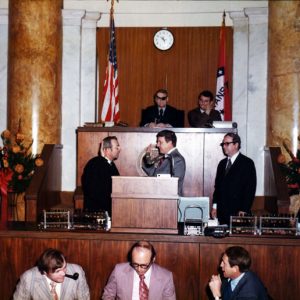 Two white men facing one another raising hands at podium, surrounded by people, with U.S., Arkansas flags