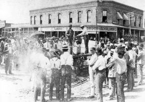 Two men on wagon with display and crowd observing, brick drugstore in background