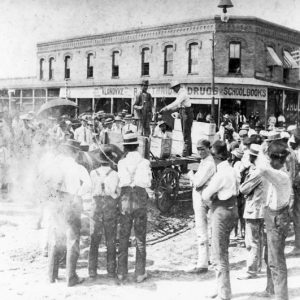 Two men on wagon with display and crowd observing, brick drugstore in background