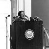 White woman speaking at podium marked "Great Seal of the State of Arkansas" with ten microphones