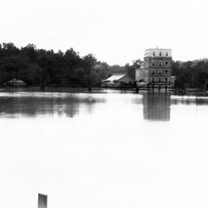 Lake with bridge and various brick buildings in background along wooded shoreline