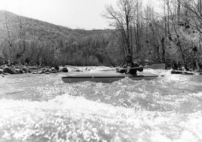 White man in kayak holding paddle across chest with river rapids, mountain ridge