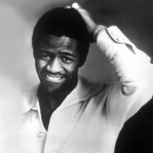 African-American man in button-up shirt smiling with his left hand on his head